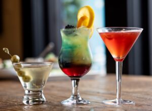 Three cocktails on a wooden table: a martini with olives, a layered blue and red drink with orange slices and a blackberry, and a red cocktail in a martini glass.