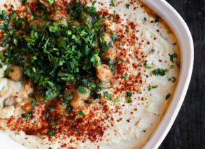 A close-up of creamy hummus topped with chopped parsley, whole chickpeas, and a generous sprinkle of red paprika, served in a white dish.