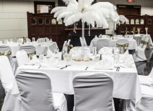 A banquet table with white tablecloth, white chair covers, and a tall centerpiece of white feathers, surrounded by similar decorated tables in an elegant indoor venue.