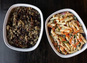 Two white dishes on a dark table: one with a rice and lentil mixture topped with caramelized onions, and one with pasta mixed with vegetables like carrots and herbs.