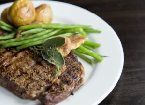 Grilled steak on a white plate, topped with herbs, served with roasted potatoes and green beans, on a dark wooden table.