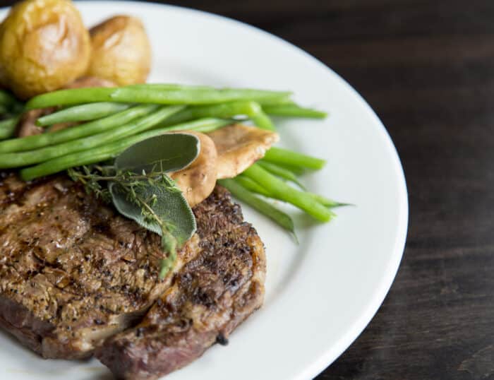 Grilled steak with fresh herbs, green beans, roasted potatoes, and mushrooms served on a white plate against a dark background.