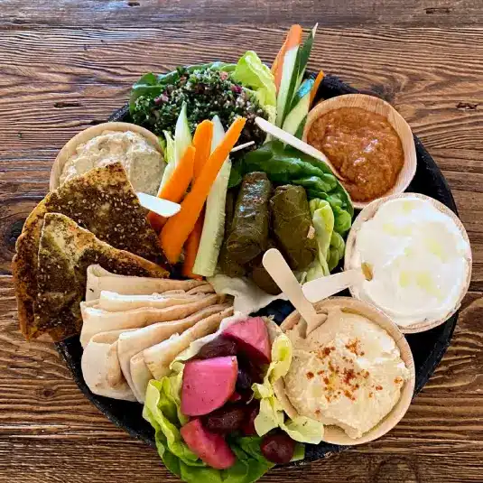 A platter with pita bread, hummus, pickled vegetables, grape leaves, fresh veggies, tabbouleh, dips, and crackers, arranged on a wooden table.